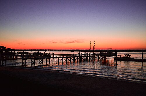 Wooden boat docks silhouetted against an orange sunset over calm sound water, flags on their poles.