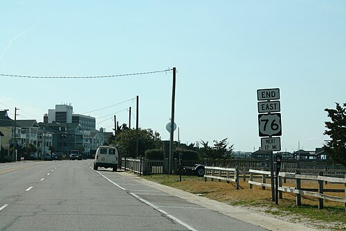 A paved two lane shore road with a wood rail fence on one side, a parked van, utility poles, a white water tower, and a beachfront hotel in the distance.