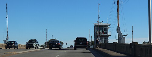 The drawbridge deck onto the island, cars crossing between concrete safety barriers and traffic gates, the bascule control tower and counterweight mechanism rising beside the roadway.