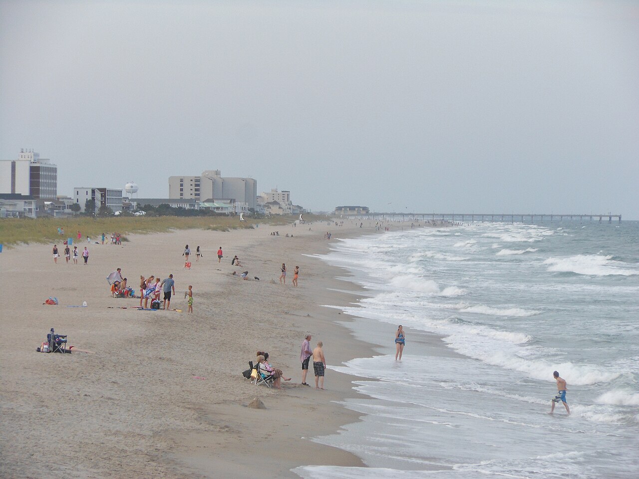 A wide sandy beach with surf rolling in, beachgoers scattered down the sand, a fishing pier reaching into the ocean a half mile off, and multi storey beachfront buildings and a water tower behind the dune line.