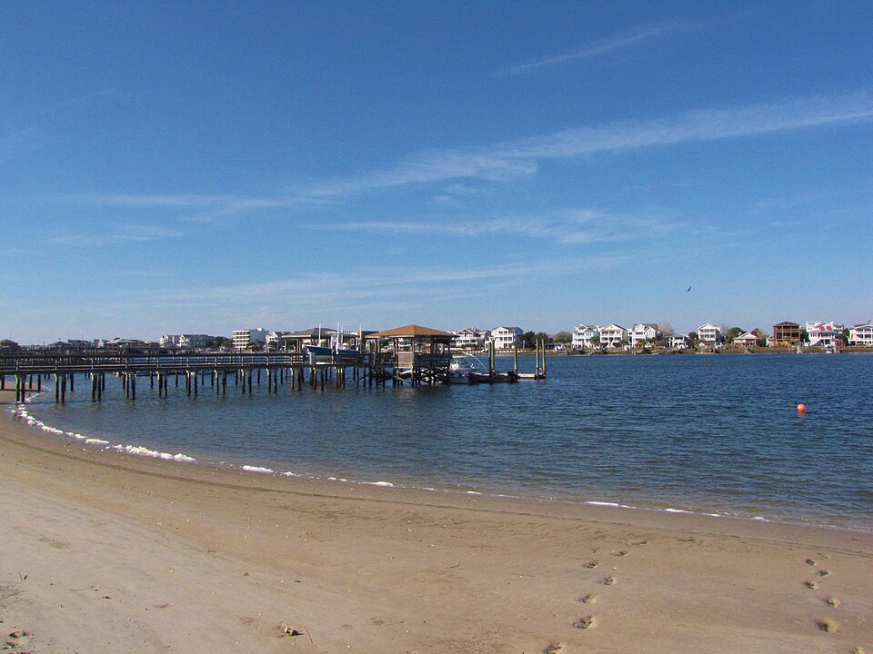 Wooden boat docks and pilings along Banks Channel, low beach houses across the water under a clear sky. The kind of Intracoastal waterfront the restaurant sits on, rather than the restaurant building itself, which is the Street View beside it.