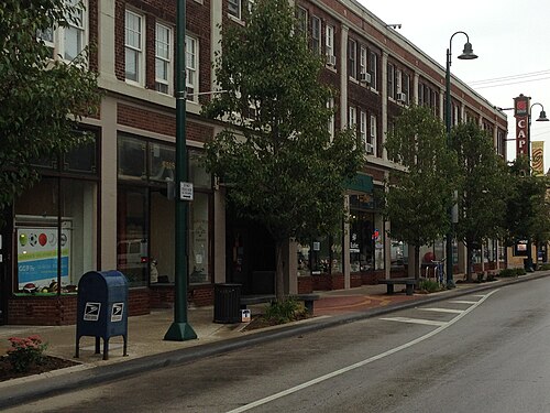 A sidewalk row of glass storefronts under young trees, benches and a mailbox along the kerb, the theatre's marquee visible down the block.