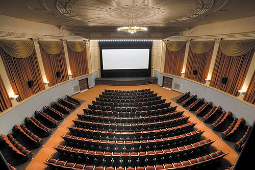 The Capitol Theatre's restored auditorium, rows of orange seats curving toward a single screen under a plaster ceiling medallion and gold curtains.