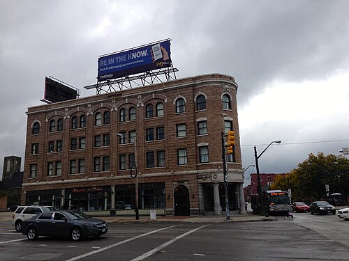 The Forest City Bank Building's curved brick corner at West 25th Street and Detroit Avenue, a restaurant under the ground floor arches and a billboard on the roof.