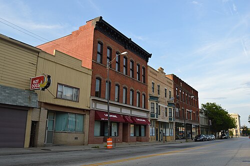 A row of two and three storey brick storefronts on Lorain Avenue, a neon hot dog sign over one shopfront and a parking meter along the curb.