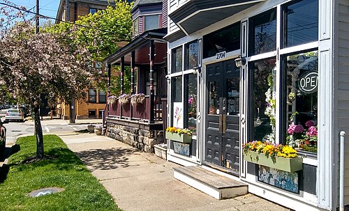 A flower shop's black storefront doors and window boxes beside a Victorian porch house, a flowering tree over the sidewalk.