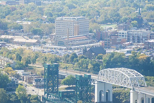 An aerial view over the market's roof and parking lot toward a lift bridge and a steel arch bridge across the Cuyahoga River, rowhouses and a church spire below.