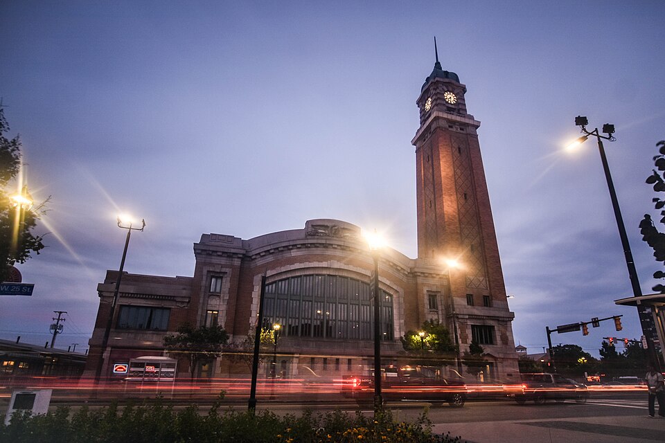 The market hall and clock tower at dusk from West 25th Street, traffic light trails crossing in front of the arched windows.