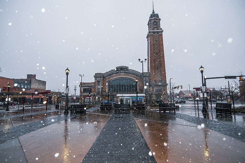 The market hall and clock tower across the paved plaza during a snowfall, benches and light poles lining the walkway to the entrance.