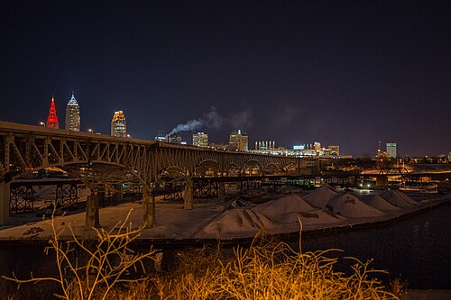 Salt storage mounds on the riverbank at night, a long bridge overhead and the lit downtown skyline behind.