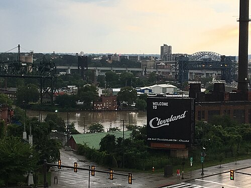 An elevated view over the river, cranes and a green roofed warehouse, with a Welcome to Cleveland sign painted on a brick building.