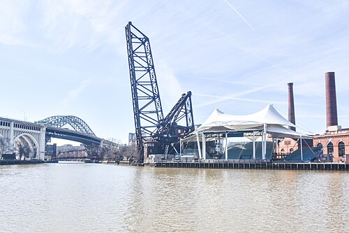 A white event tent and old brick smokestacks on the riverbank, with the raised railroad lift bridge and the Detroit-Superior Bridge behind them.
