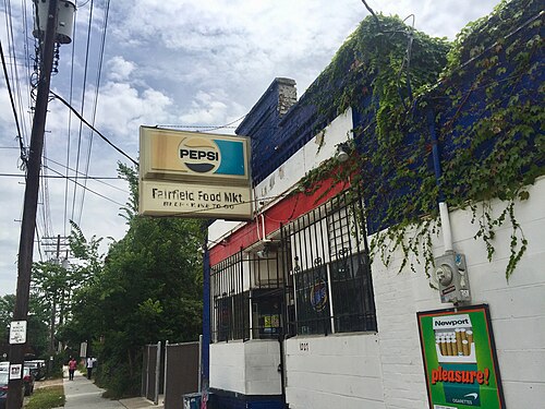 A corner store painted white, red and blue, ivy climbing the upper wall, a Pepsi sign over hand lettered Fairfield Food Mkt lettering and iron bars on the windows.