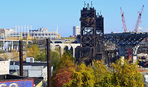 A steel lift bridge and construction cranes over the Cuyahoga, Progressive Field and the downtown skyline visible beyond warehouse roofs and smokestacks.