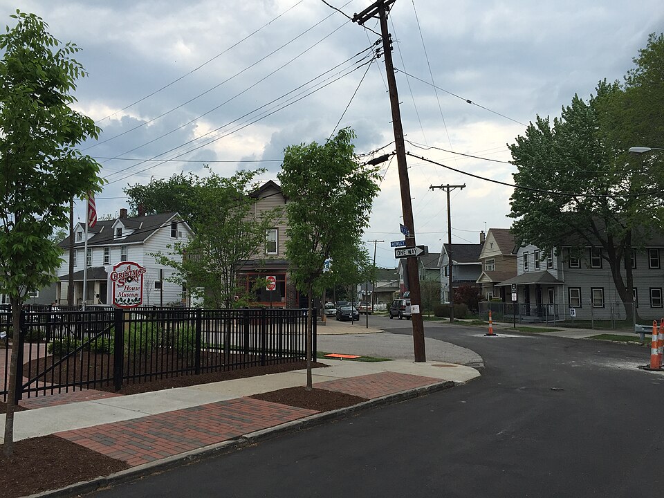 The A Christmas Story House Museum sign mounted on a black iron fence, the restored house behind young trees and a brick paver walkway, wood frame houses continuing up the block.