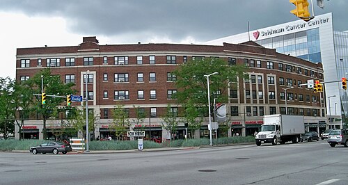 A curved red brick building with ground floor shops following the street corner, the glass fronted Seidman Cancer Center behind it.