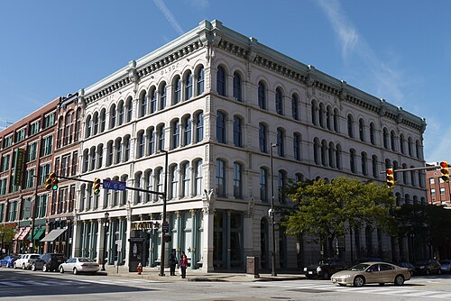 The Hoyt Block's pale carved stone corner at West 6th and St. Clair, arched windows stacked over cast iron ground floor storefronts, traffic signals at the intersection.