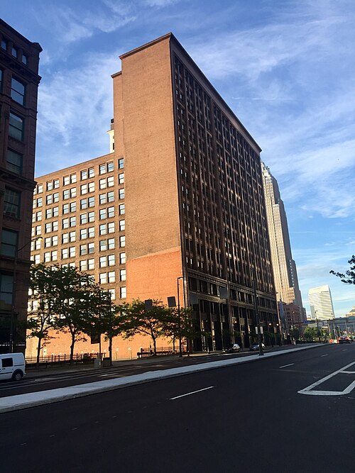 The Rockefeller Building's narrow steel framed tower rising above a lower brick warehouse on Superior Avenue, taller glass towers visible behind it.