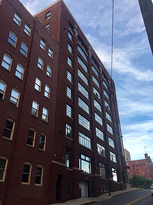 A tall red brick warehouse building converted to apartments, unbroken window bays rising from base to cornice on a sloped street.
