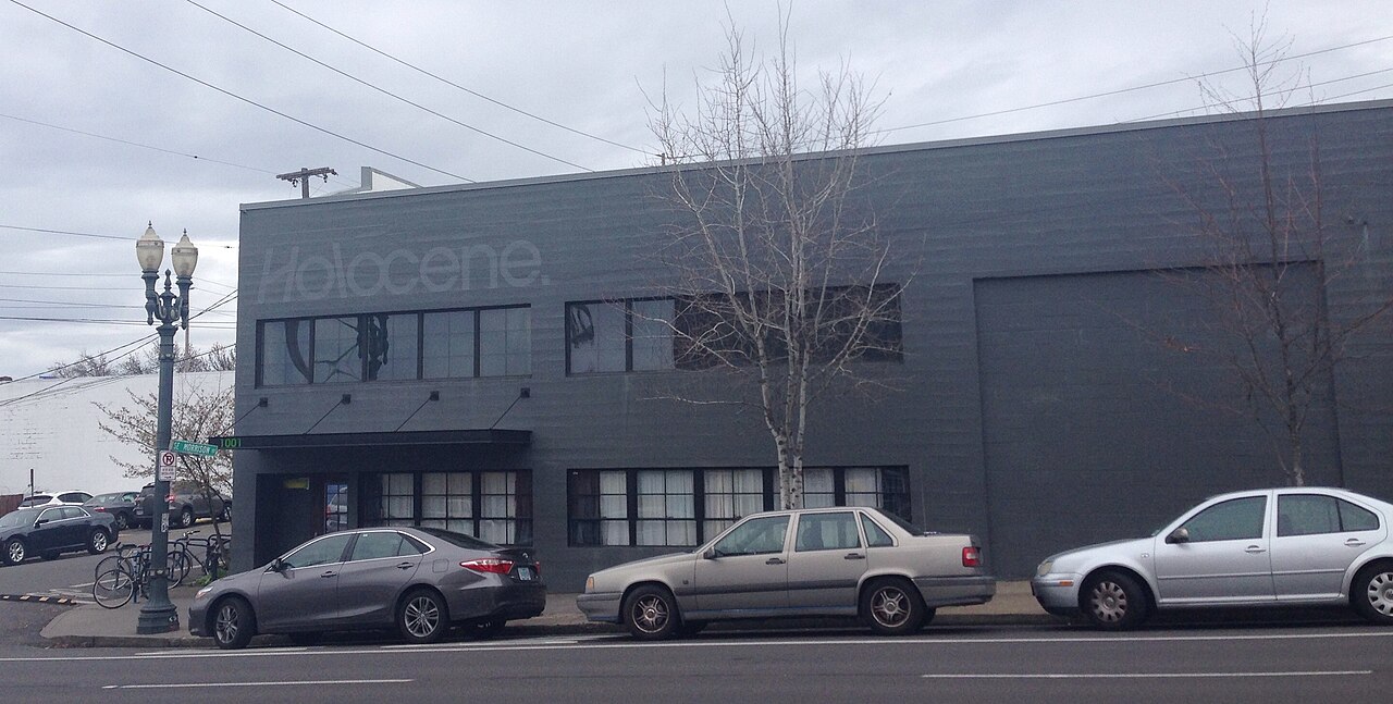 A dark painted warehouse building with a roll up loading door and upper floor windows, cars parked along the curb.