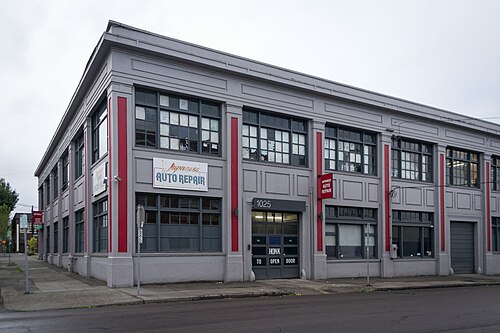 A two storey grey painted garage building from 1921 with red pilasters, tall multi pane windows and a roll up bay door at one end.