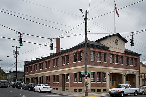 A two storey brick fire station on a corner, built in 1927, with an American flag and traffic signals overhead.