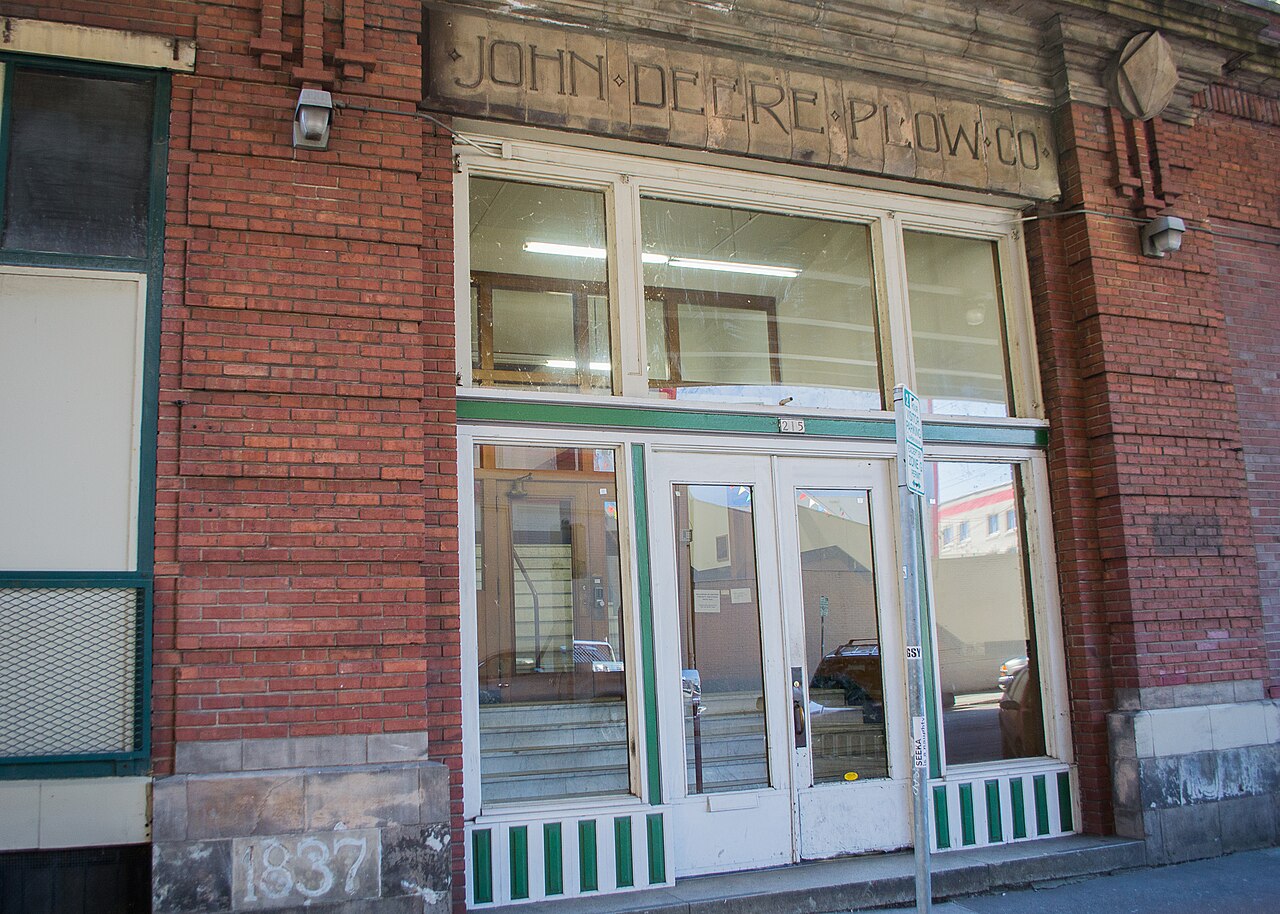 The brick entrance of the John Deere Plow Company Building at 215 SE Morrison Street, its 1911 carved sign over green trimmed transom windows and doors.