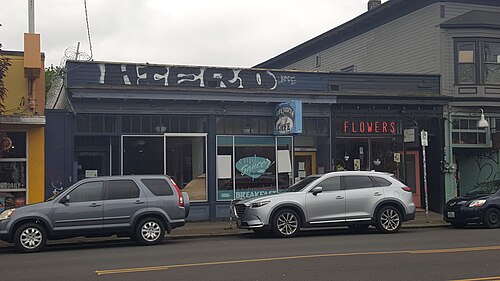 A one storey shopfront row painted dark blue and yellow, a lit FLOWERS sign, and cars parked along the curb.