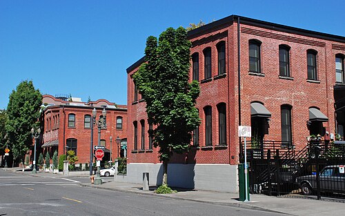 Two matching two storey red brick depot buildings facing each other across NW 11th Avenue, round arched windows and a street tree screening part of the nearer building's corner.