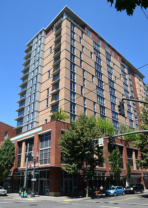 A tan and red brick apartment tower over ground floor retail at NW 12th and Lovejoy, balconies stacked up its rounded corner.