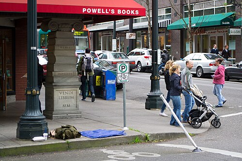 The Powell's Books awning and a carved stone column reading Vende Librum on the sidewalk at NW 11th and Couch, pedestrians and parked cars along the curb.