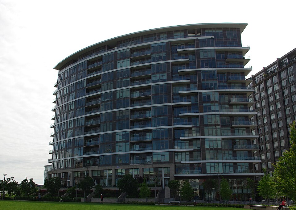 A curved glass and brick apartment tower rising above The Fields Park's lawn, its floors ringed with balconies.