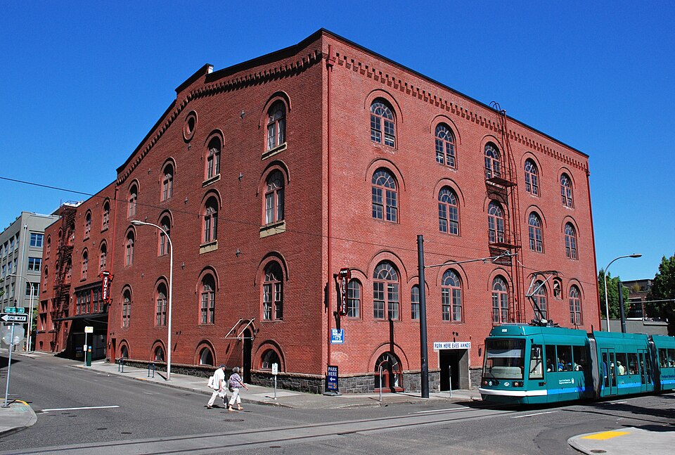 A four storey red brick warehouse with round arched windows on every floor, a fire escape zigzagging up one corner, and a Portland Streetcar passing at street level.