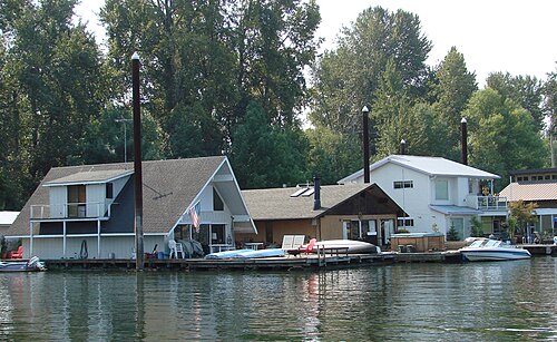 Houseboats moored along the Willamette riverbank, trees along the shore behind them.