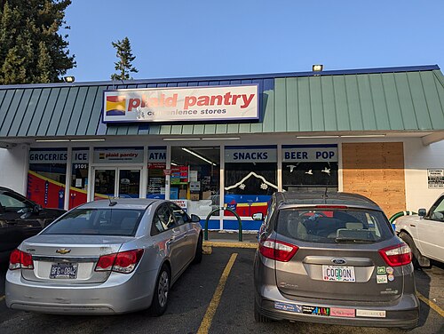 The Plaid Pantry convenience store's green metal roofline, plywood over one window and cars nosed in at the curb.