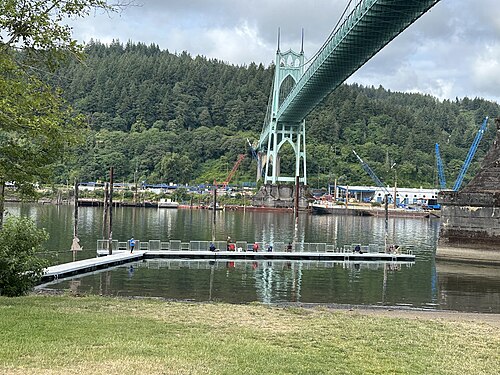 A floating dock in Cathedral Park, a few anglers along the rail, the bridge's east tower and concrete piers rising straight out of the water behind it.