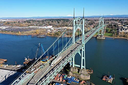A high aerial view of the bridge with Mount Hood and Mount St. Helens on the horizon, cranes and barges working the river below.