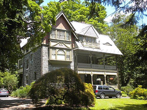 A stone and shingle Victorian house with a two tier wraparound porch, red trim gables and a parked SUV in the drive.