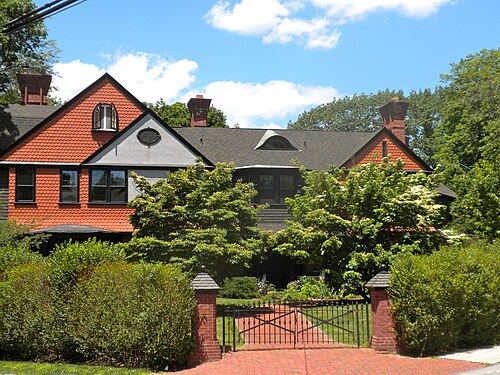 A red fish scale shingle gable over rough stone, brick gateposts and an iron gate at the drive, hedges along the sidewalk.