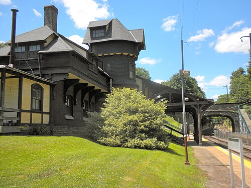 A dark shingled train station with a corner turret and deep eaves, a covered platform running alongside the tracks.