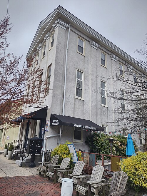 A plain stucco corner building with tall windows and a black awning over the entrance, wooden chairs out front.