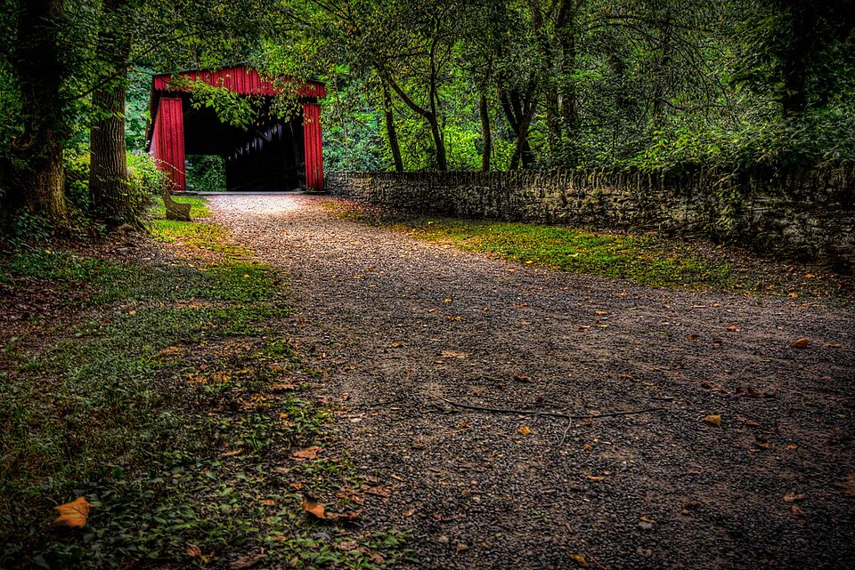 A red wooden covered bridge over the Wissahickon Creek on Forbidden Drive, a gravel path leading up to it between stone retaining walls.