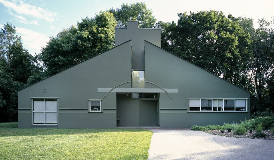A low grey house with its whole front cut into one triangular gable, a chimney breaking through the peak and an arched lintel over the recessed door.