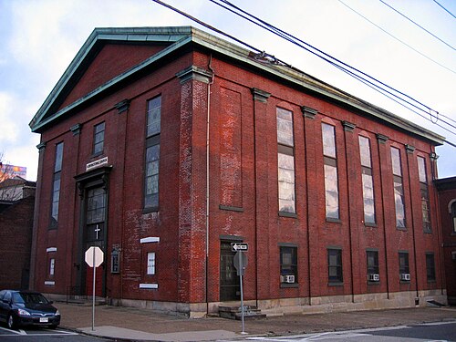 A red brick church at a corner, arched stained glass windows down each side, a copper edged pediment roof and a small cross over the main door.