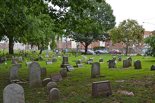 Headstones under mature trees at Palmer Cemetery, small American flags on some graves, brick rowhouses visible past the cemetery fence.