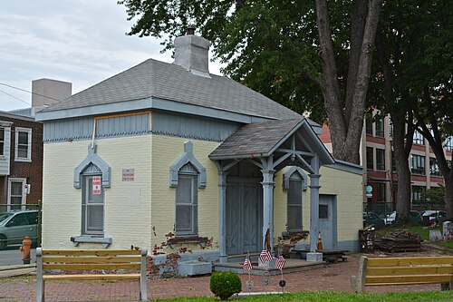 The cemetery's pale yellow Victorian gatehouse, a steep slate roof, gothic arched window surrounds and a small covered porch with American flags at the steps.