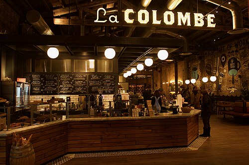 The counter inside La Colombe's roastery on Frankford Avenue, exposed timber roof trusses, hanging globe lights and a lit marquee sign over the register.