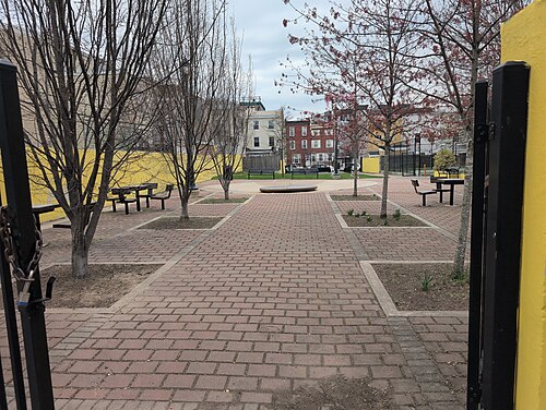 A small brick paved pocket park, young trees in cutouts, benches along the sides and brick rowhouses behind a painted yellow perimeter wall.