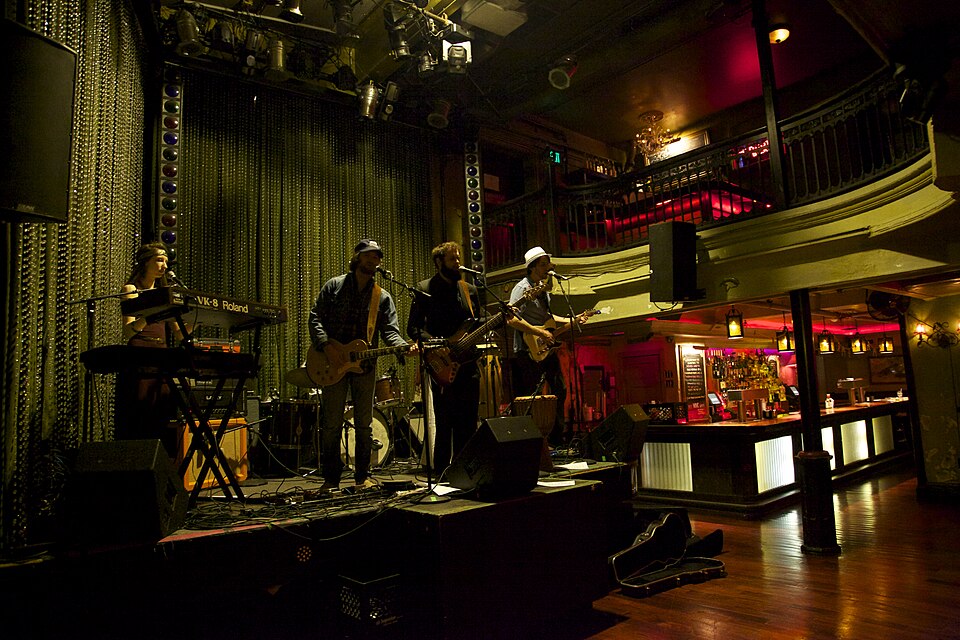 The interior of Johnny Brenda's during a show, musicians on the small stage under the venue's two tier balcony, the ground floor bar and its string lights to the right.
