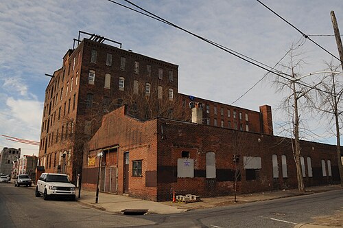 A six storey red brick mill building wrapping a street corner, boarded upper windows and loading doors along the ground floor base.